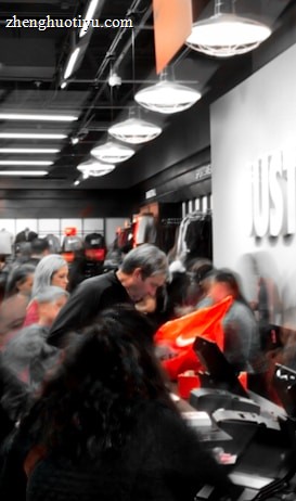 A busy retail store with numerous people examining products and clothing. The store appears to be selling athletic wear, with the phrase 'Just Do It.' prominently displayed on a wall alongside a recognizable orange logo. The interior is well-lit with modern lighting, shelves filled with merchandise, and a counter where people are gathered, possibly checking out or asking questions.