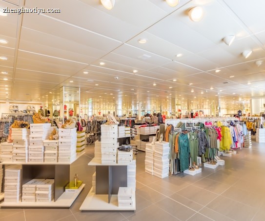 A brightly lit, spacious retail clothing store with rows of neatly arranged clothing racks and shelves filled with folded garments. The store has high ceilings with numerous spotlights and a modern, minimalist design. Multiple stacks of white shoe boxes are prominently displayed on tables throughout the space.