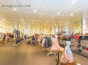 A spacious retail store with bright lighting and neatly arranged clothing and footwear displays. Mannequins wearing children's outfits are prominently featured in the foreground, while various clothes and shoes are organized on racks and shelves throughout the store. People are seen browsing and shopping in the background, adding a lively atmosphere to the setting.