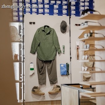 A retail display inside a store featuring outdoor or casual clothing and accessories arranged on a white board. Items include a green long-sleeve shirt, gray pants, a pair of green and beige shoes, a green hat, sunglasses, a pair of white socks, a small green item, and a blue pouch. In the background, shelves are stacked with blue and white shoe boxes.
