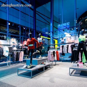 A spacious, modern clothing store with mannequins displayed on raised platforms. The mannequins are dressed in athletic wear, including leggings, sports bras, and jackets. Large glass windows provide an outside view, letting in natural light. Clothing racks filled with various apparel items, including leggings, shorts, and tops, line the walls. The overall setting is clean and organized.
