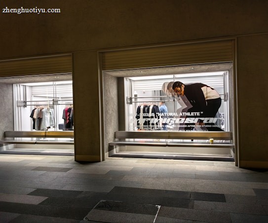 A store display with two large glass windows, featuring clothing racks with various items such as shirts and jackets. There is a poster of a person bending down, with text 'Natural Athlete' across it. The surrounding area includes a tiled walkway.
