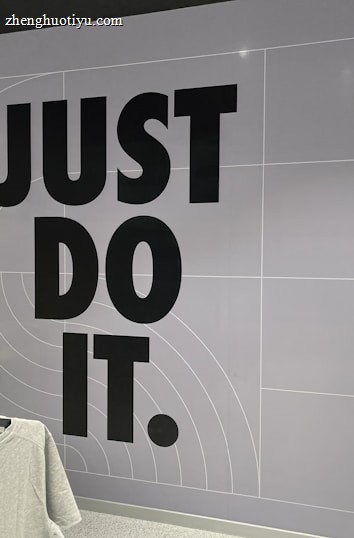 The image features a retail store interior with a prominent slogan 'JUST DO IT' in large black letters on a tiled wall. There is a clothing rack in the foreground with various T-shirts displayed on hangers. The wall has a grid pattern with subtle circular lines. To the right, a sign indicates fitting rooms, and a person is partially visible. The store is well-lit with modern fixtures.