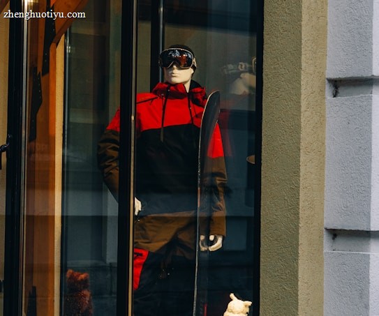 A storefront display features a mannequin dressed in a red and black ski outfit, complete with goggles and holding a snowboard. The scene is set against a snowy background with decorative logs and artificial snow, creating a winter sports theme.